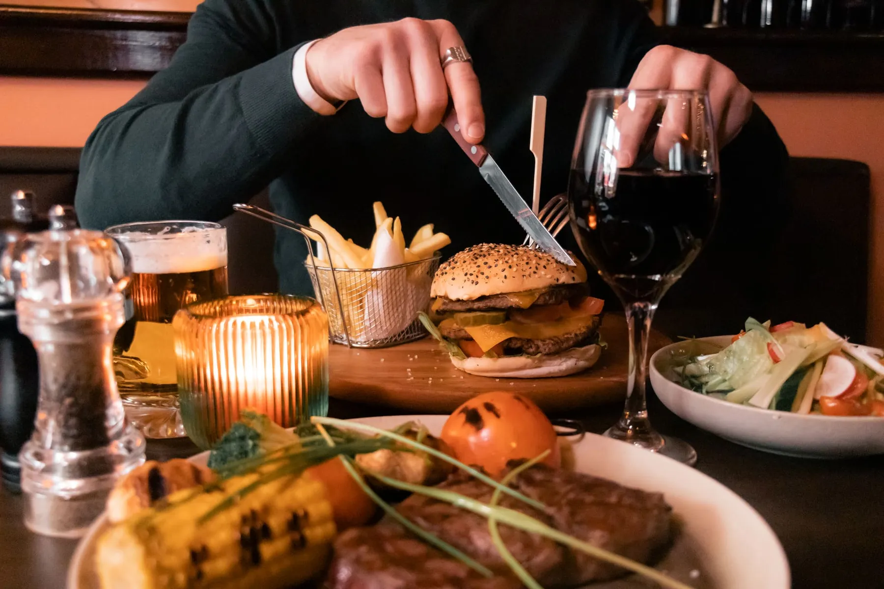 Een gast snijdt bij kaarslicht in een burger, met rode wijn, friet en salade op tafel bij La Stego in Amsterdam